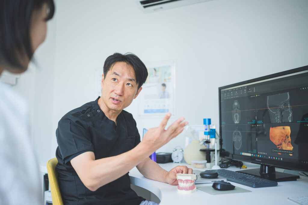 愛媛インプラントクリニックかまくら歯科にて、歯科医師が歯科用CT（3次元エックス線診断装置）の立体画像と顎模型を用いて、患者様に親知らずの抜歯計画を説明している様子。神経や血管の位置を精密に把握し、安全性を重視したカウンセリングを行っている風景。