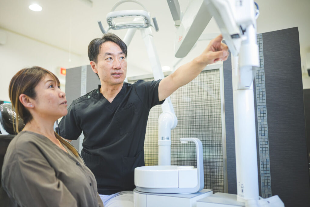 愛媛インプラントクリニックかまくら歯科にて、歯科医師が患者様にインプラント治療のカウンセリングを行っている様子。診療ユニットに座る患者様に対し、モニターや設備を指し示しながら、治療計画や手順について丁寧に説明しています。愛媛県松山市でインプラントの不安を解消し、納得のいく歯科診療を提供するワンシーンです。