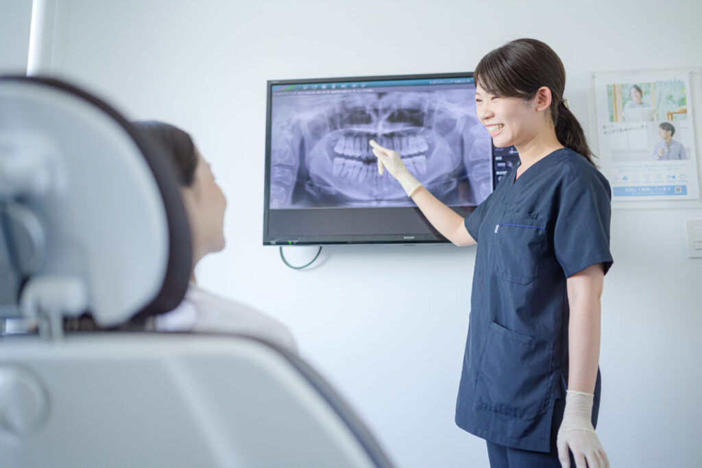 愛媛インプラントクリニックかまくら歯科にて、歯科衛生士がパノラマレントゲン画像を用いて患者様に丁寧なカウンセリングを行っている様子。大型モニターで歯の状態を指し示しながら、インプラント治療や口腔内の現状について分かりやすく説明しています。