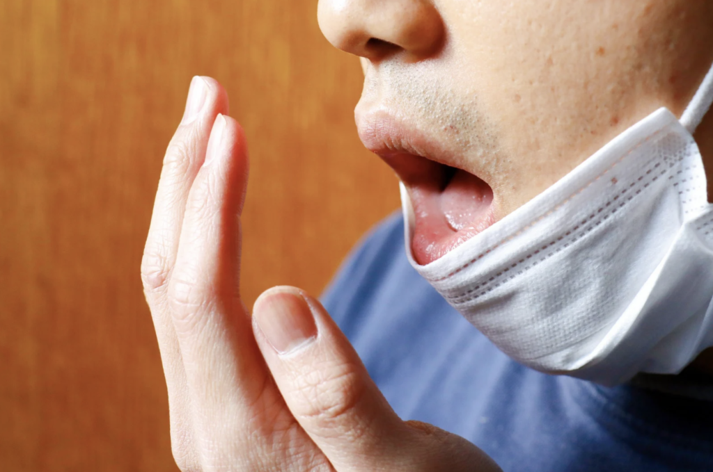 愛媛インプラントクリニックかまくら歯科が提唱する口臭セルフチェックと対策。マスクを外して自分の息のニオイを気にする男性の様子。愛媛県松山市近郊で口臭の原因となる歯周病や膿栓、舌苔などの口腔トラブルを専門的に改善するためのイメージ画像。