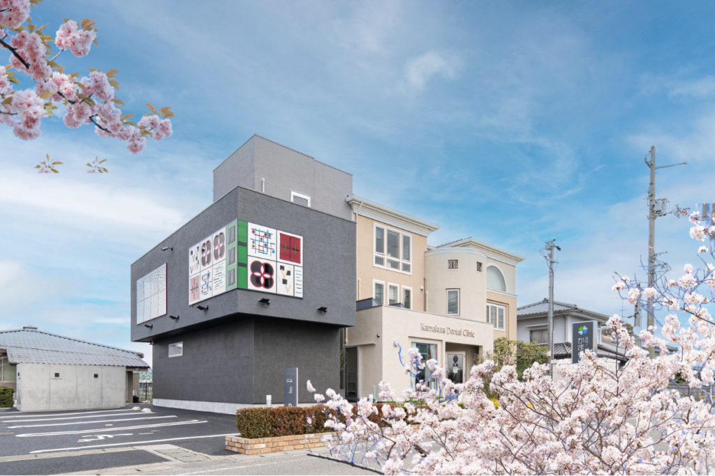 愛媛県伊予郡松前町にある「愛媛インプラントクリニックかまくら歯科」の外観。青空の下、満開の桜に彩られたモダンで清潔感のある医院建物。広い駐車場を完備し、松山市近郊からも車で通いやすいバリアフリー設計。インプラント治療の専門性を象徴するような、デザイン性の高いアートが施された壁面が特徴。