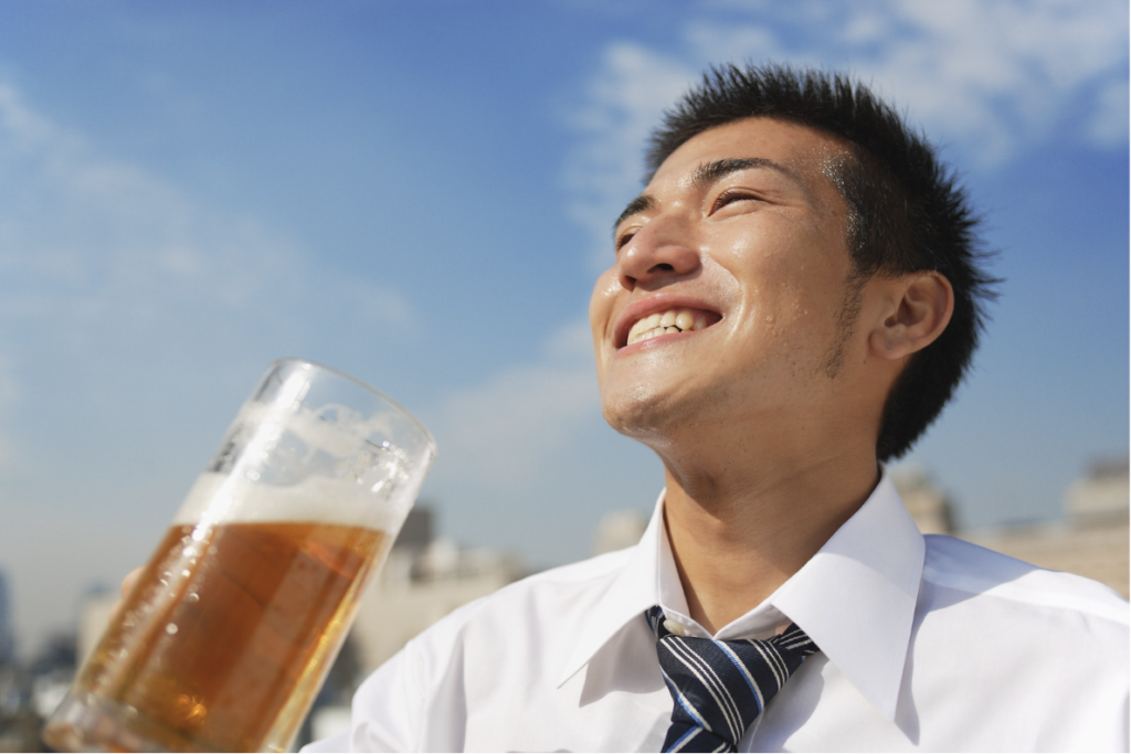 青空の下、白いワイシャツとストライプのネクタイ姿の男性が、冷たいビールの入ったグラスを片手に爽やかな笑顔を浮かべている様子。愛媛のかまくら歯科では、インプラント治療を通じて、冷たい飲み物や食事を心から楽しめる健康な歯と豊かな日常をサポートしています。
